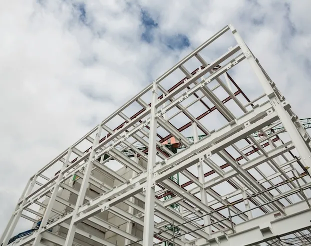 A white metal structure of a building under construction, seen from below, against a cloudy sky